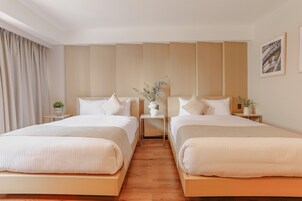 A hotel room with two beds, a wooden floor, a large window with curtains, and a small table with a plant.