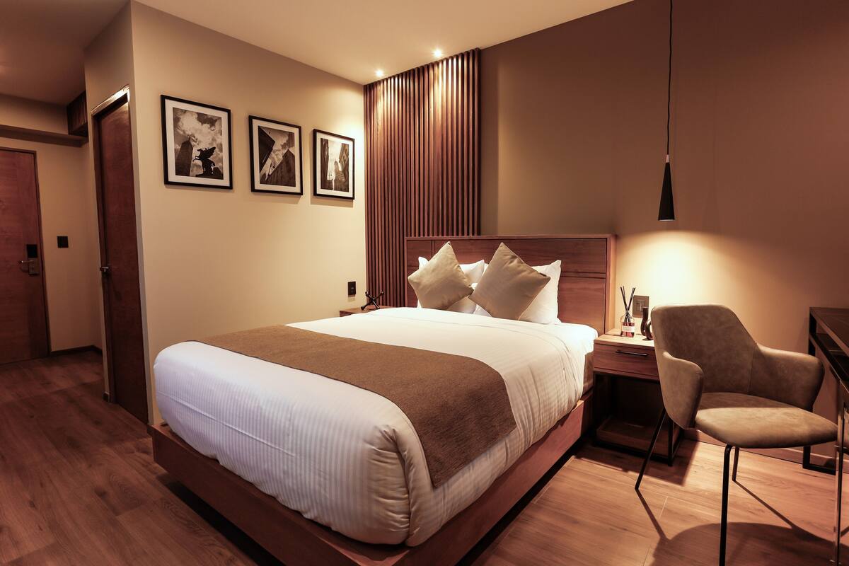 A hotel room with a large bed, a desk with a chair, and a wall-mounted lamp.