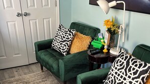 Ein Zimmer mit zwei grünen Sofas, eines mit einem schwarz-weißen Kissen und das andere mit einem goldenen. Im Hintergrund eine weiße Tür und ein gerahmtes Bild an der Wand.