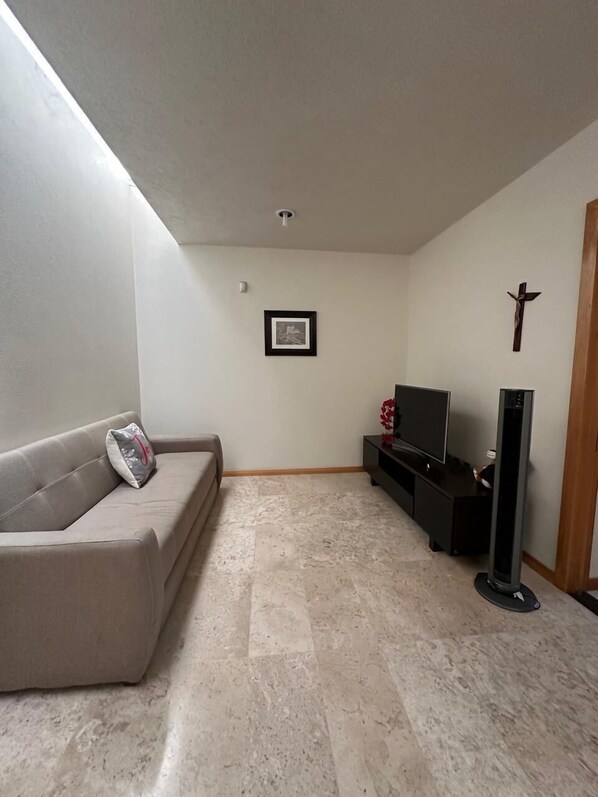 Una sala de estar con un sofá color beige, un cuadro en la pared, un televisor y un objeto alto y negro en el suelo.