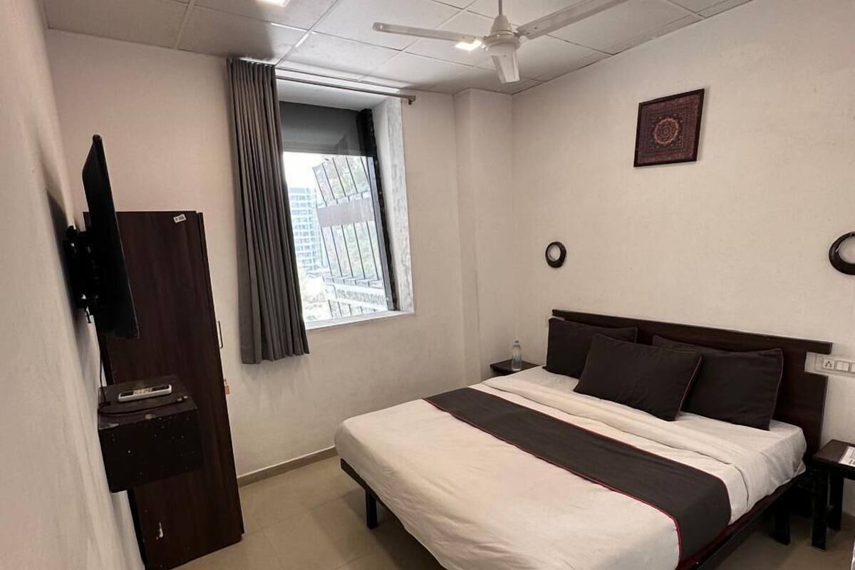 A hotel room with a bed, a ceiling fan, a window with curtains, a wall-mounted TV, and a small table.