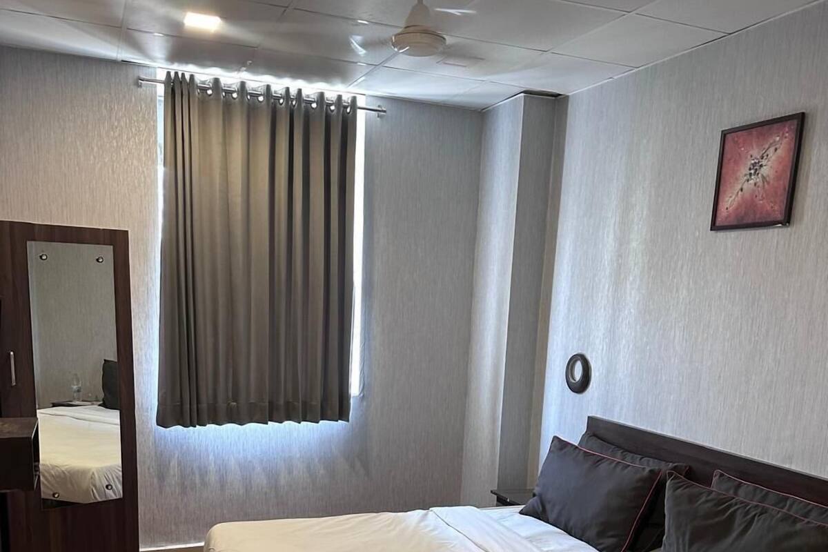 A hotel room with a large bed, a ceiling fan, a window with curtains, a mirror, and a painting on the wall.