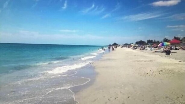 Een strand met helderblauw water en een zandstrand. Er staan parasols en mensen op het strand.