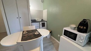 Een kleine keuken met een magnetron, een fornuisje en een spoelbak. Er staat een witte tafel met een tafelkleed met een patroon, en een kopje. De muren zijn groen en er is een witte deur op de achtergrond.