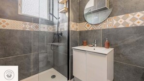 A modern bathroom with a glass-enclosed shower, a white sink with a round mirror, and patterned tile walls. - Le duplex inverse, Bollaert, Fac, gare, 2pers (Lens)