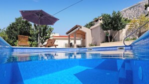 A swimming pool with a tiled edge, a wooden deck, and a covered seating area with a table and chairs. - Stunning home in Bast (Bast)