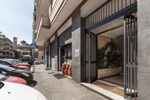 Un edificio con una entrada moderna, un estacionamiento con coches y una calle visible con otros edificios.