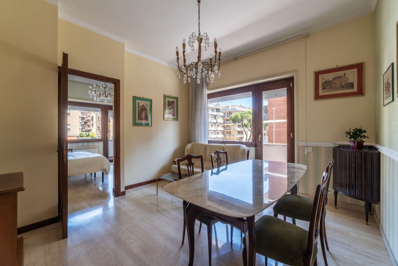 Un comedor con una mesa de mármol, sillas de madera, una araña y vistas a edificios a través de amplios ventanales.