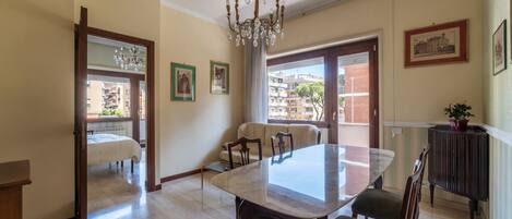 Un comedor con una mesa de mármol, sillas de madera, una araña y vistas a edificios a través de amplios ventanales.