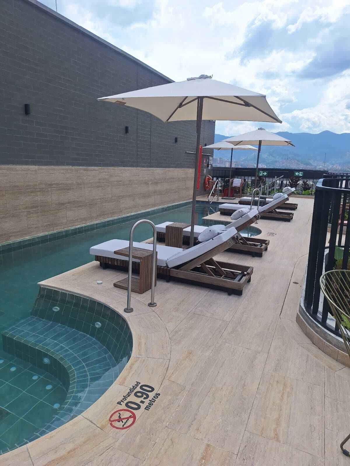 A rooftop pool area with lounge chairs and umbrellas, a sign indicating a depth of 0-90 meters, and a view of the city and mountains in the background.