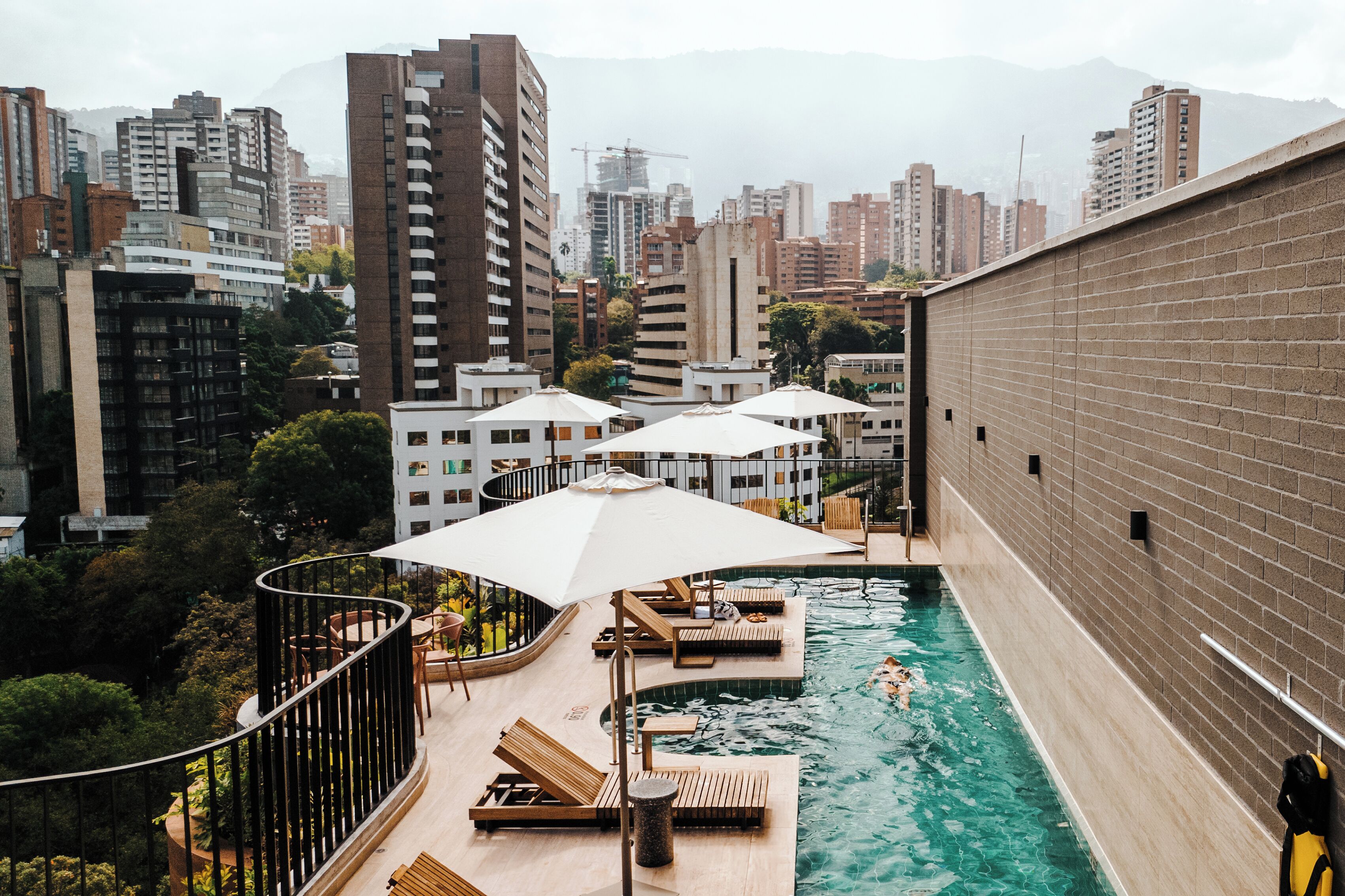 Une piscine sur le toit, dotée de chaises longues et de parasols, entourée d’immeubles de grande hauteur et de verdure.