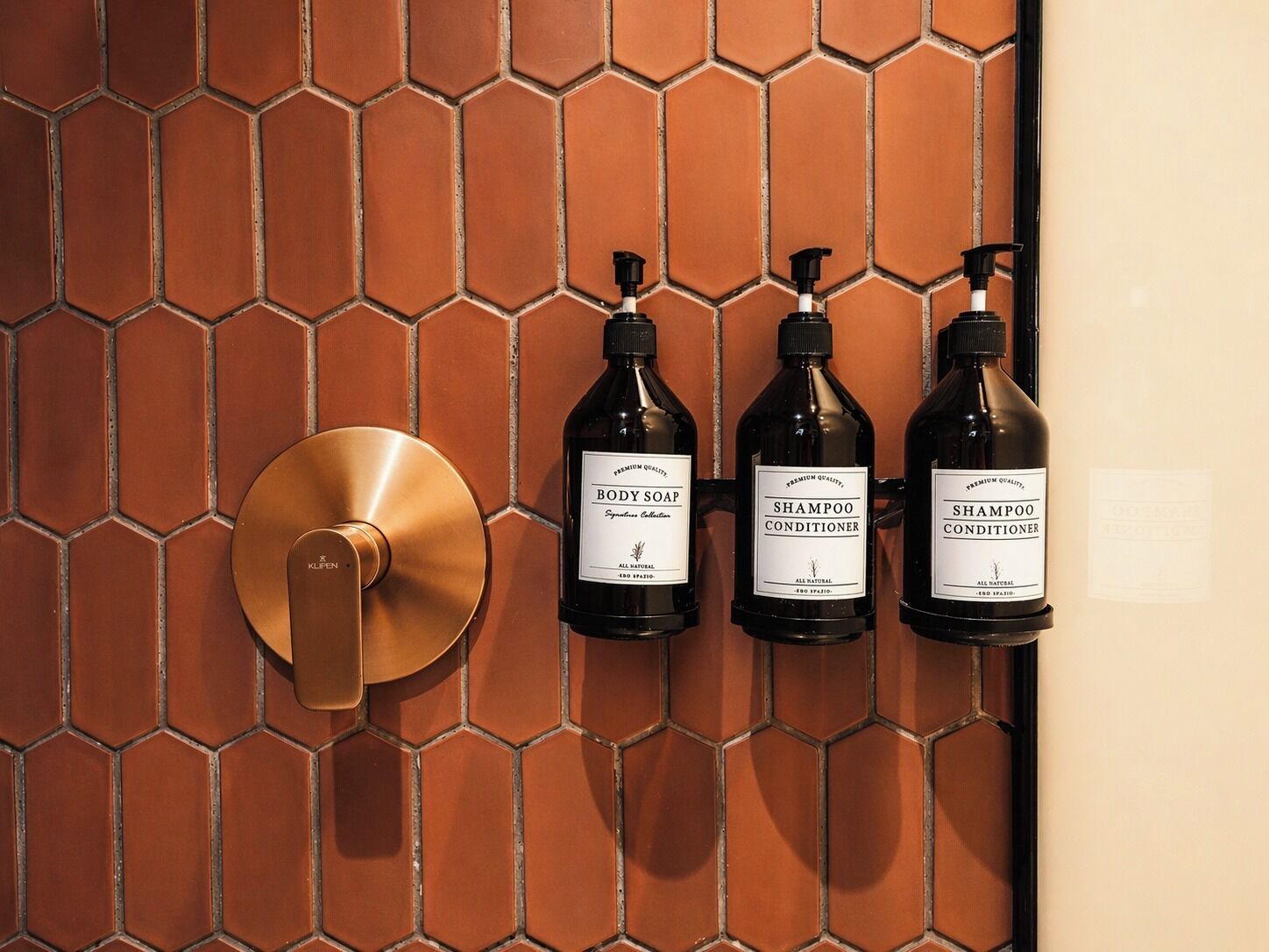 Une salle de bain avec un mur recouvert de carreaux hexagonaux, une poignée de porte en laiton et trois flacons de gel douche, de shampoing et d’après-shampoing.