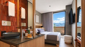 A hotel room with a wooden vanity, a bed, a large window with a mountain view, and a flat-screen TV mounted on the wall. - Heiss Hotel By Jalo (Medellín)