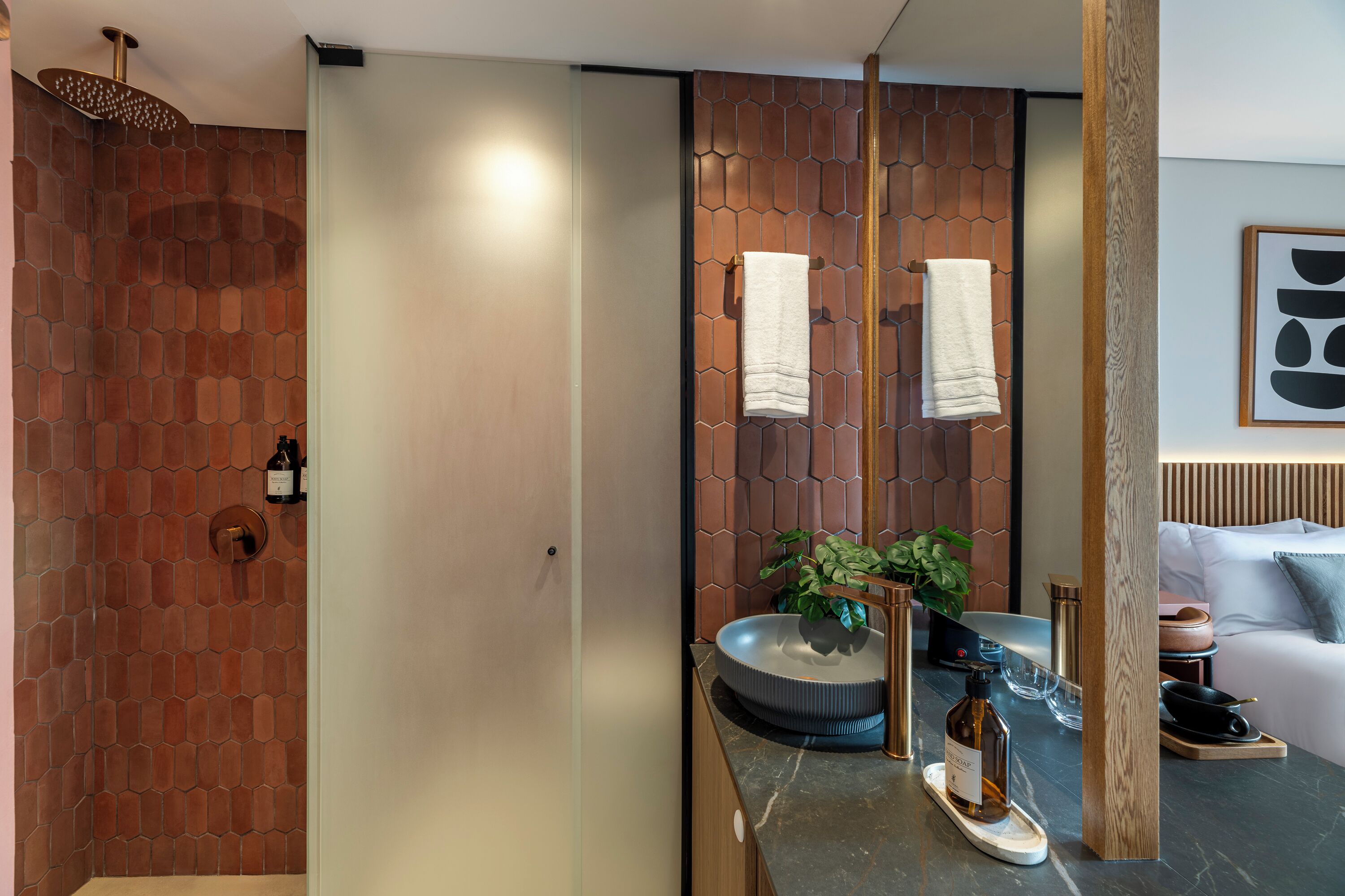 A modern bathroom with a brick wall, a large mirror, a sink with a bowl, a bottle of olive oil, and a potted plant.