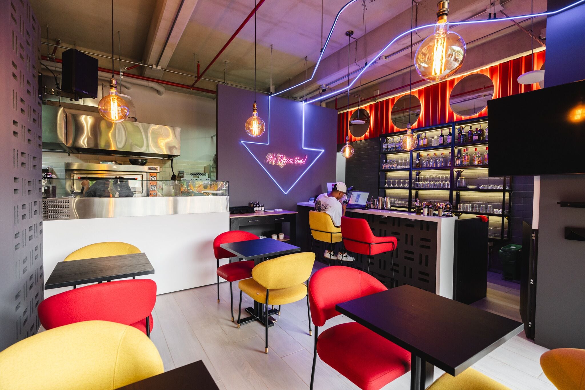 Zona de bar moderna con un letrero de neón, barra y asientos con sillas rojas y amarillas.