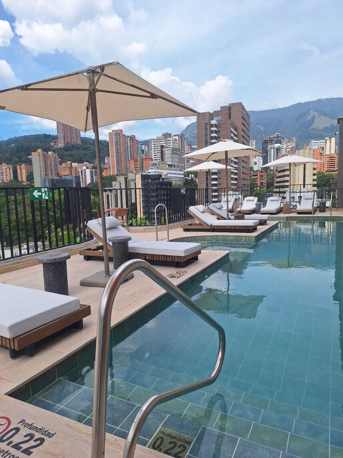 An outdoor swimming pool with lounge chairs and umbrellas, surrounded by high-rise buildings and a mountainous backdrop.