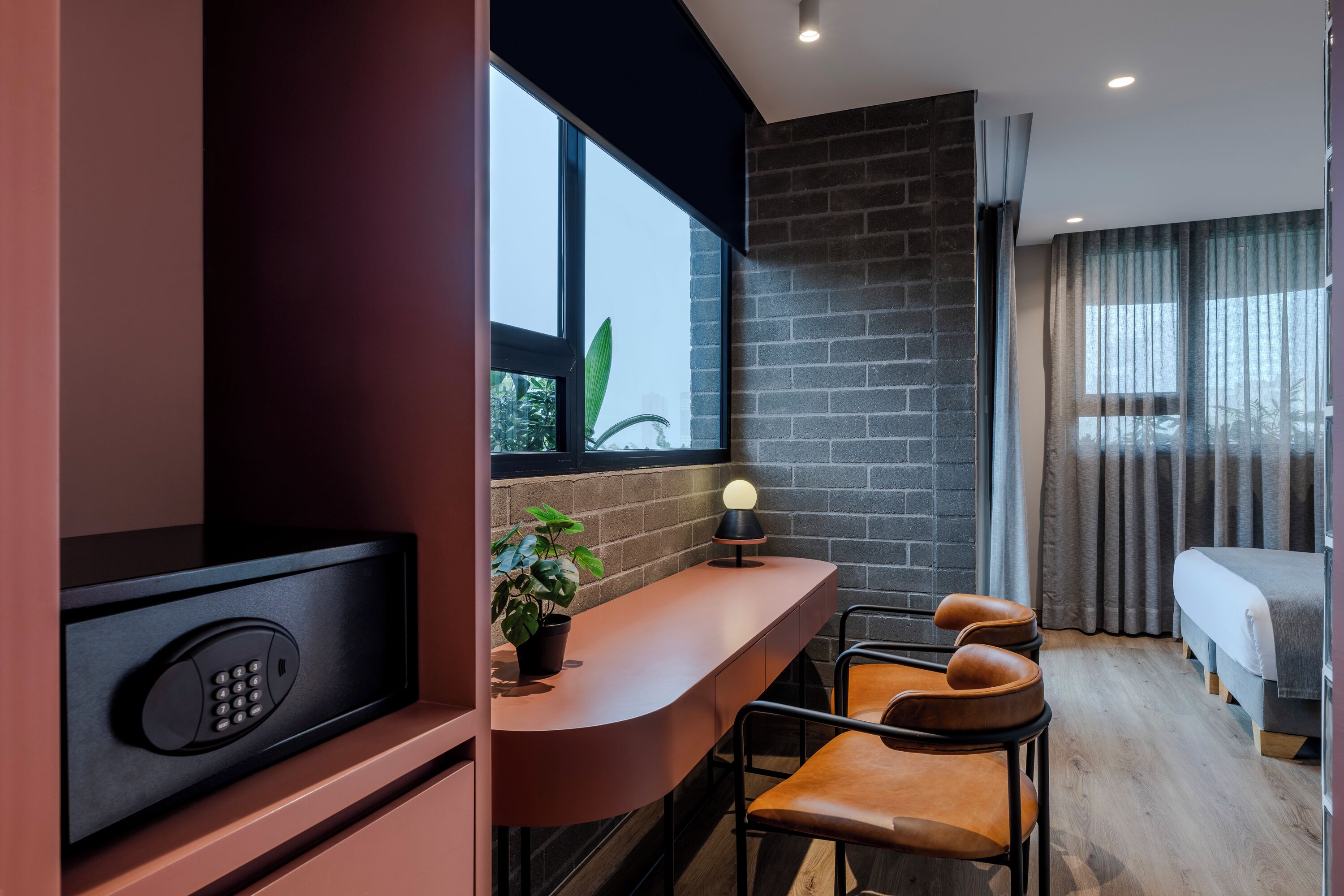 A modern hotel room with a built-in desk, a potted plant, a lamp, a bed, and a window with curtains.