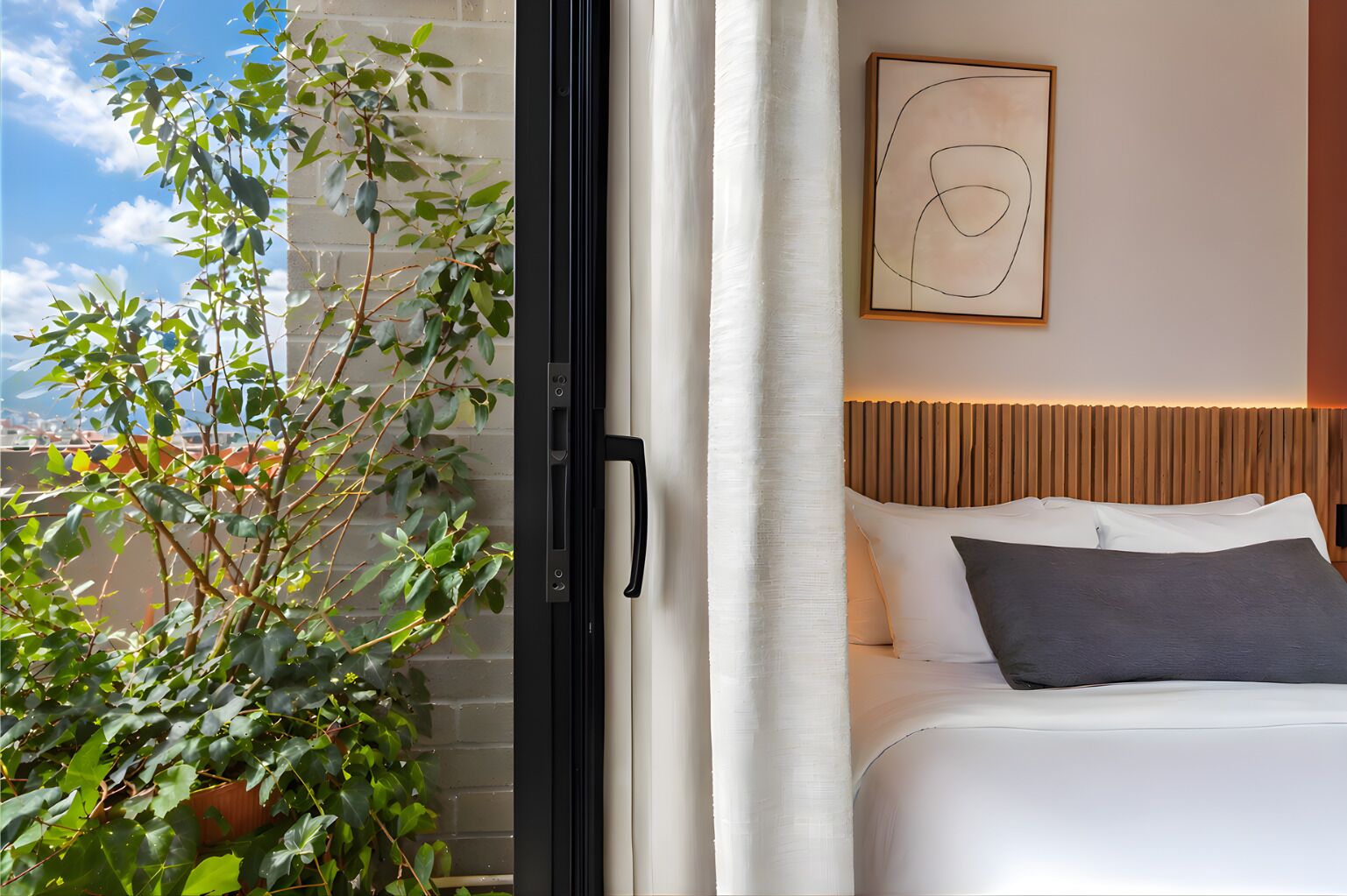 A hotel room with a bed, a window with a view of a brick wall and greenery, and a modern abstract painting on the wall.