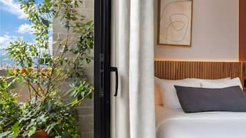 A hotel room with a bed, a window with a view of a brick wall and greenery, and a modern abstract painting on the wall.