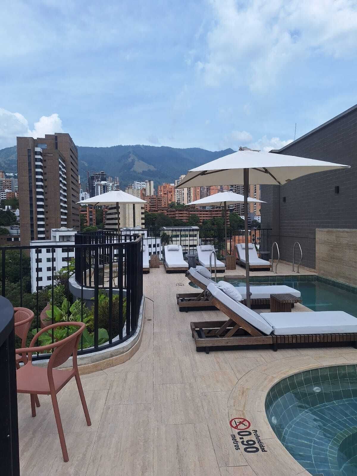 A rooftop pool area with lounge chairs, umbrellas, and a view of the city and mountains.