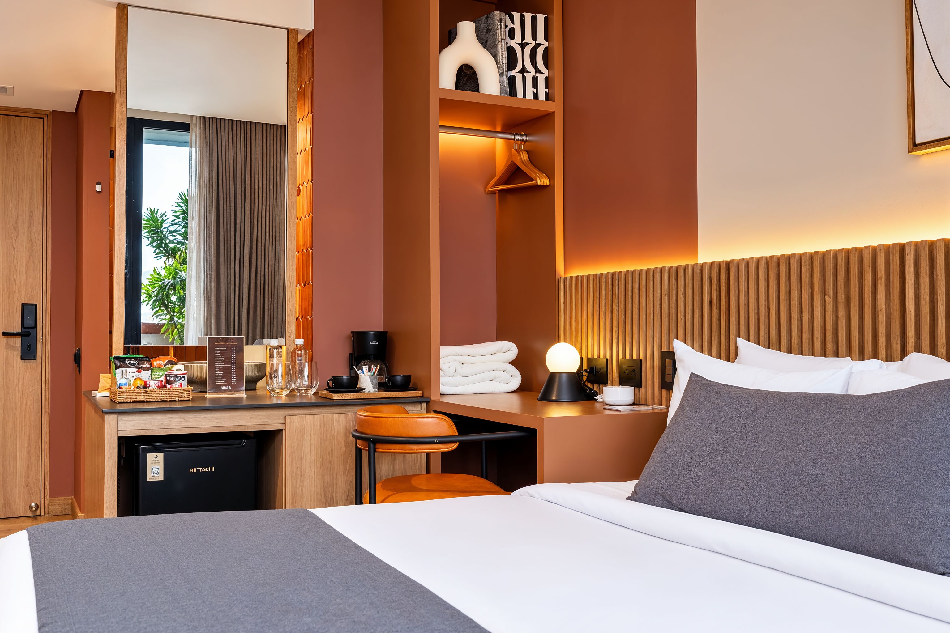 A hotel room with a bed, a desk with a coffee machine, a minibar, and a view of greenery outside.