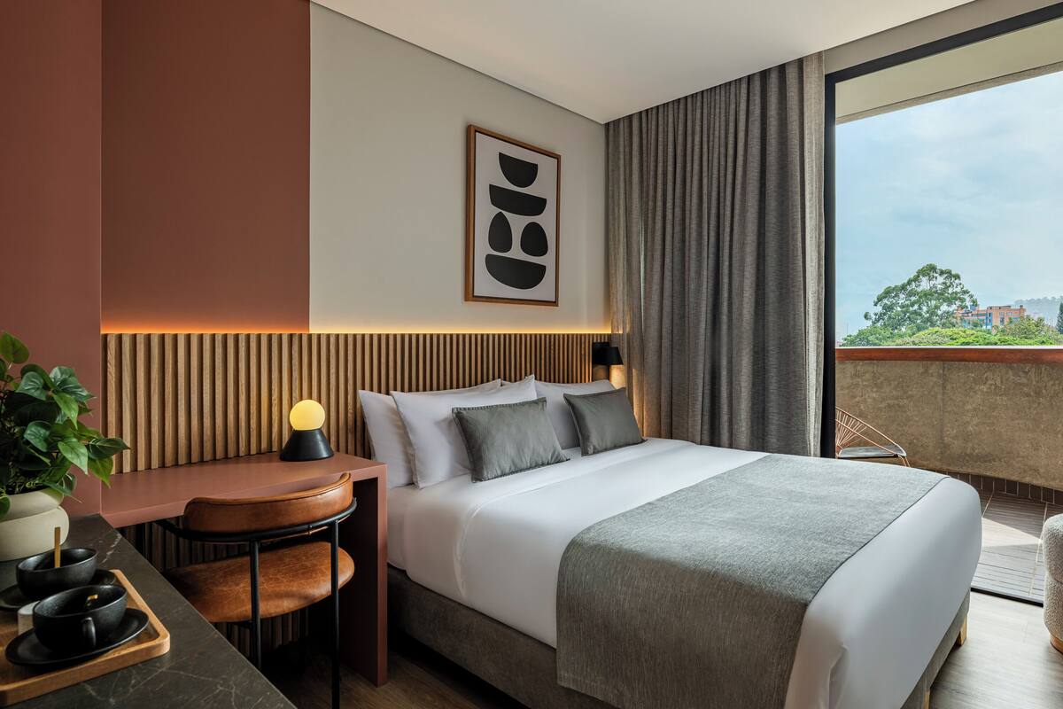 A modern hotel room with a large bed, a desk with a lamp, a potted plant, and a city view.