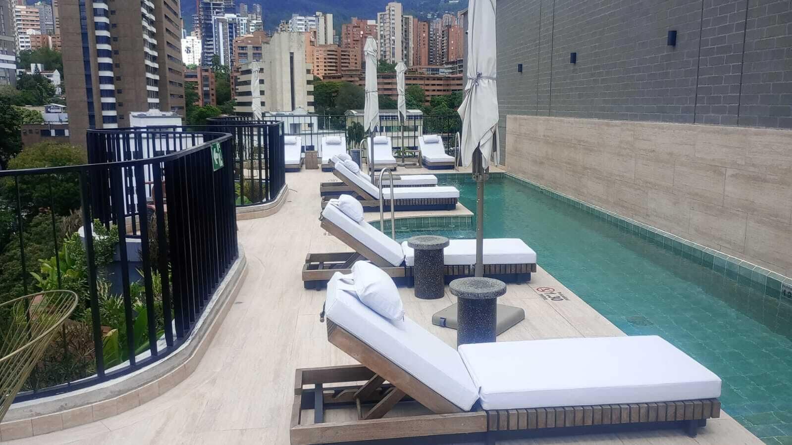 Un área de piscina en la azotea con sillones y tumbonas, rodeada de un paisaje urbano.
