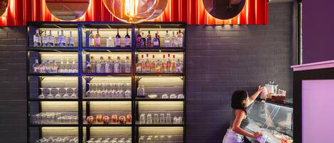A bar area with shelves of bottles and glasses, a woman reaching for a bottle, and a neon light fixture.
