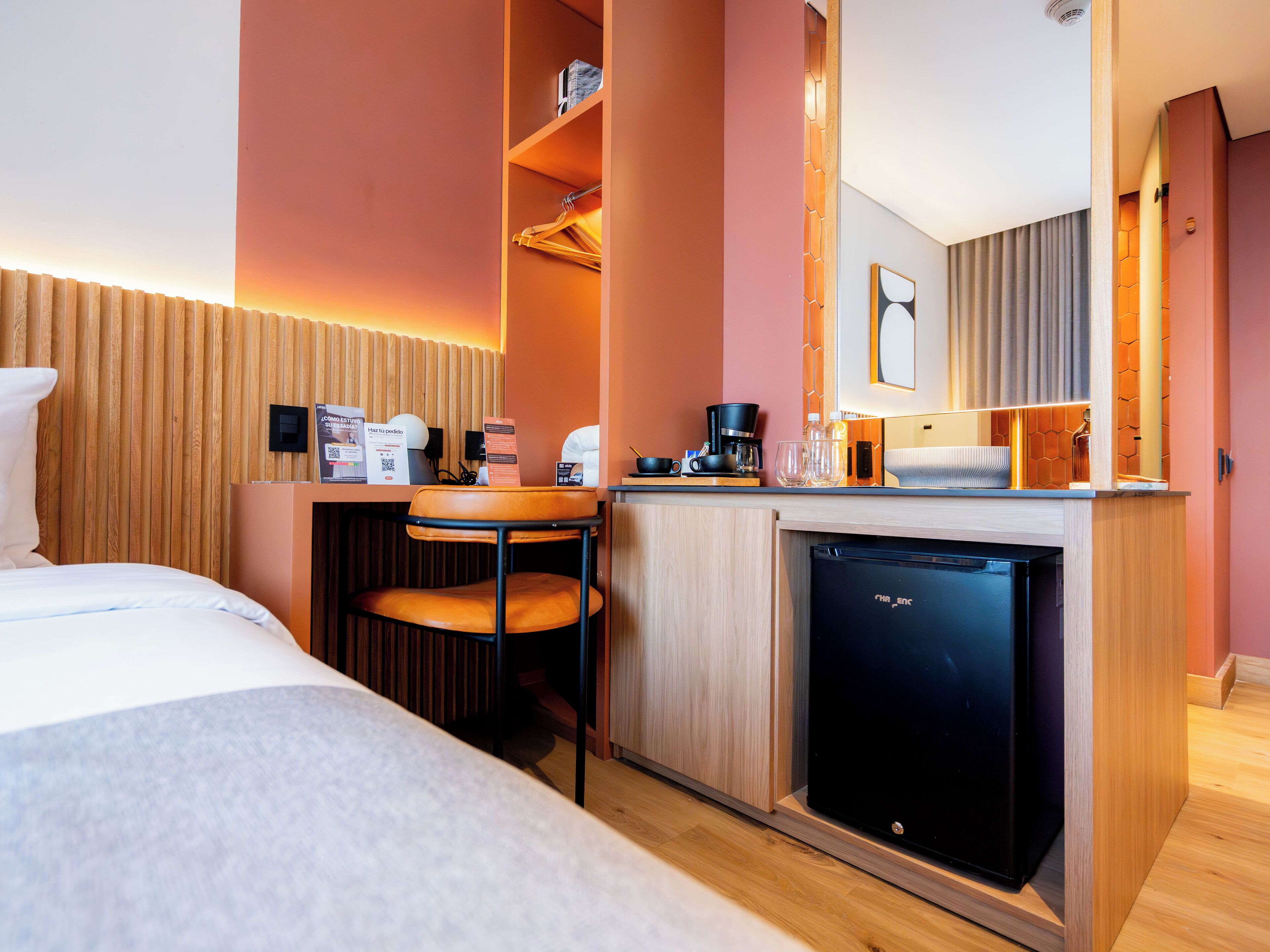 A modern hotel room with a bed, a small kitchenette, a chair, and a compact refrigerator.