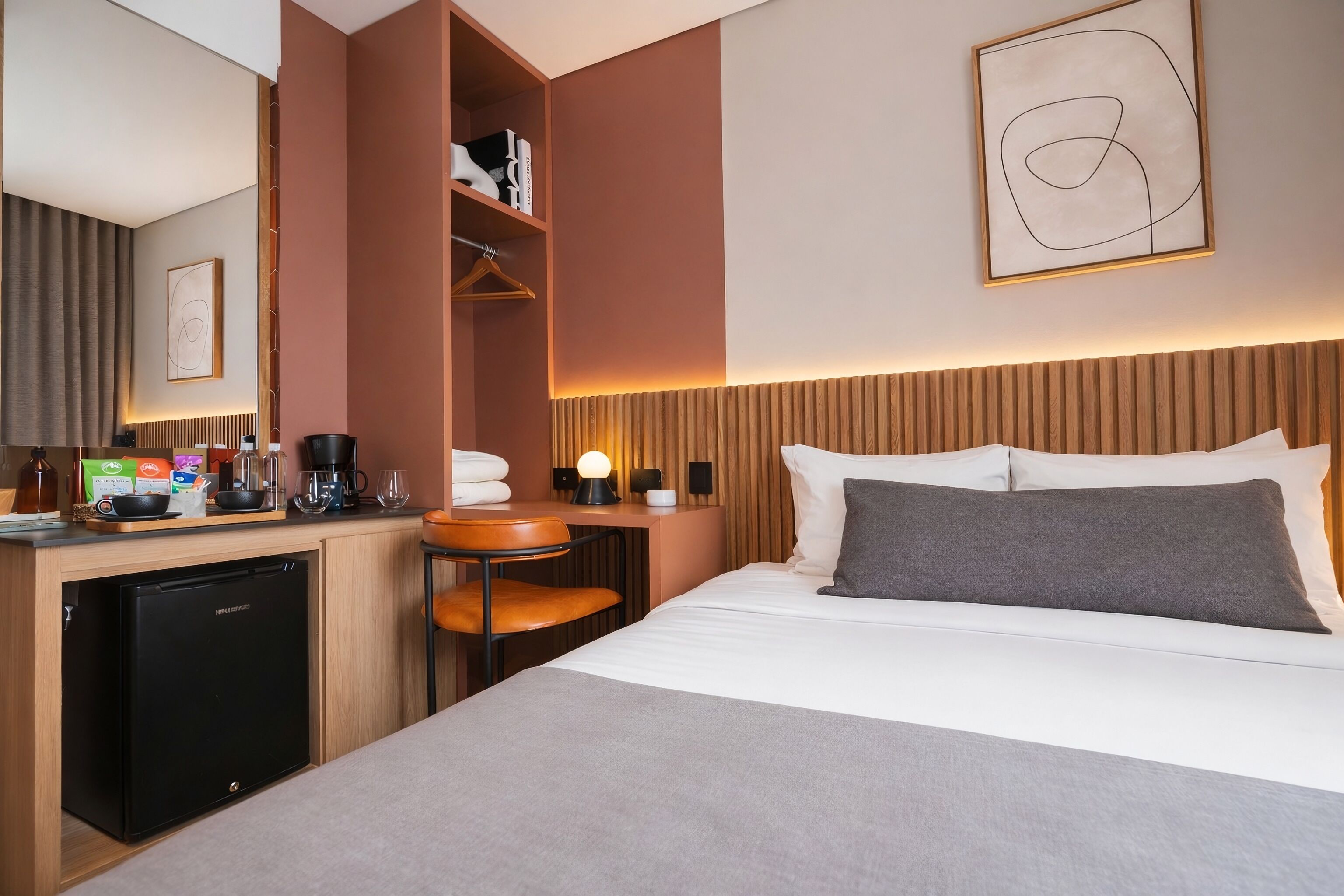 A hotel room with a bed, bedside table, a desk with a coffee machine, a mini fridge, and a wardrobe.