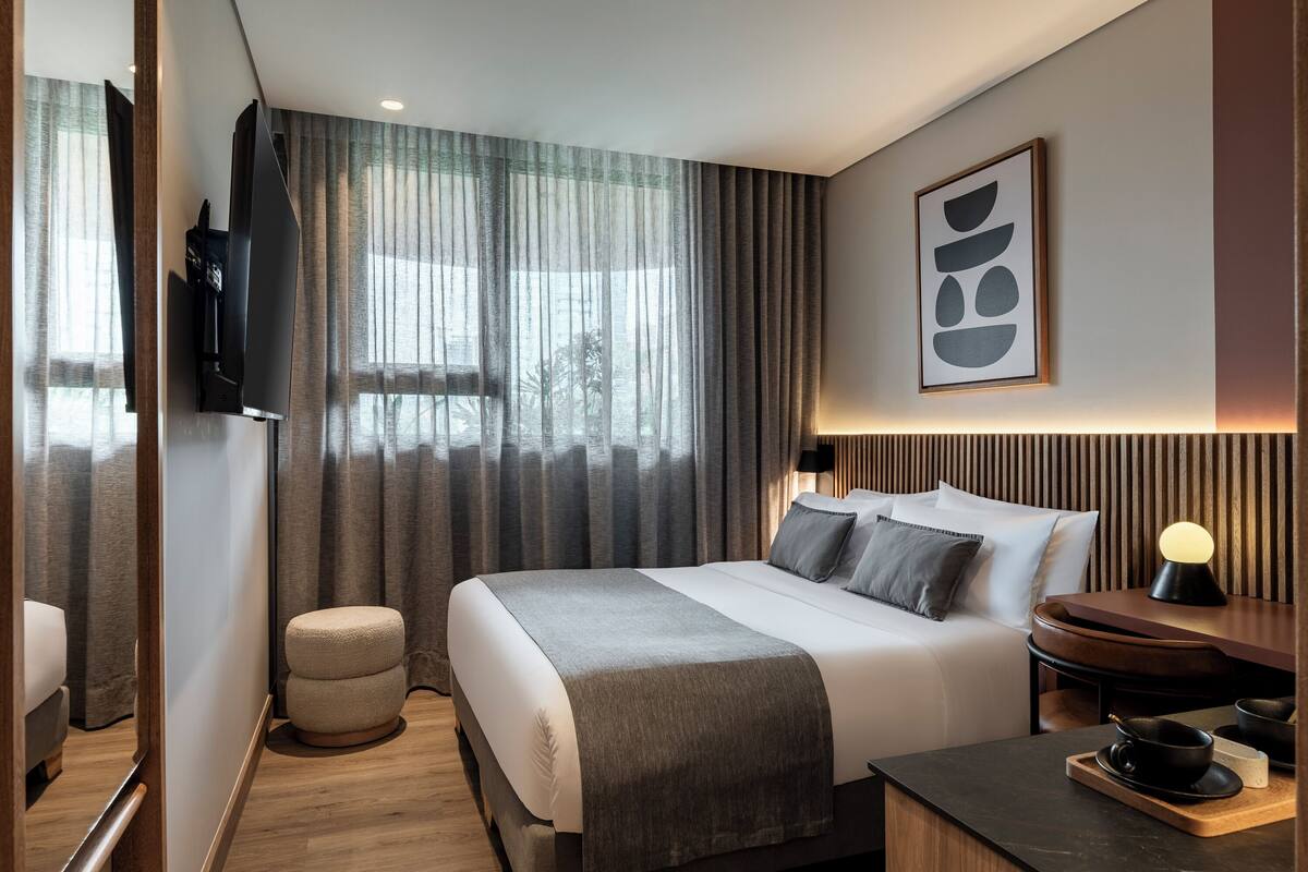 A modern hotel room with a large bed, a bedside table with a lamp, a small stool, and a TV mounted on the wall.
