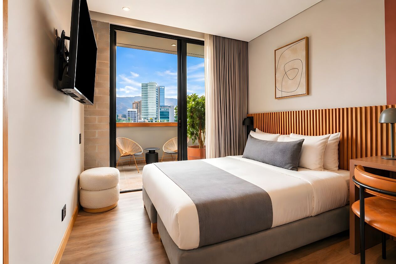 A modern hotel room with a large bed, a TV mounted on the wall, a balcony with a view of the city, and a potted plant.