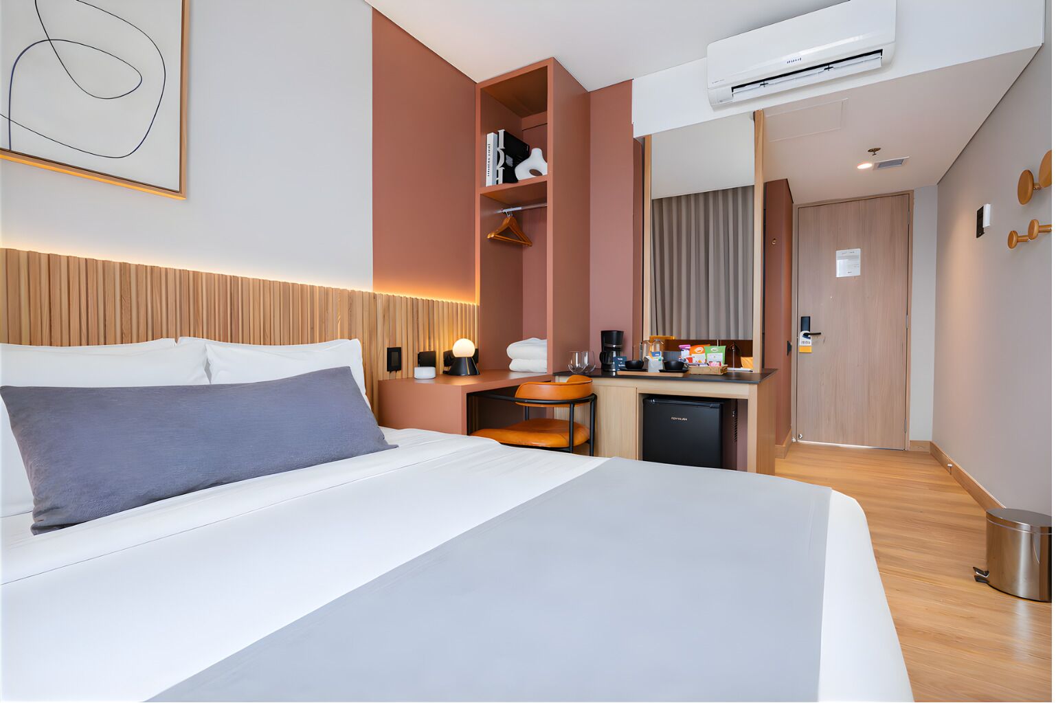 A hotel room with a bed, bedside table, lamp, and a kitchenette area.