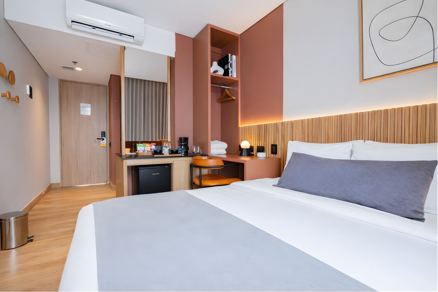 Quarto de hotel com cama, escrivaninha, mini-geladeira, cafeteira e luminária pendente.
