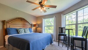 Une chambre avec un grand lit, un ventilateur de plafond, deux fenêtres et une vue sur la verdure.