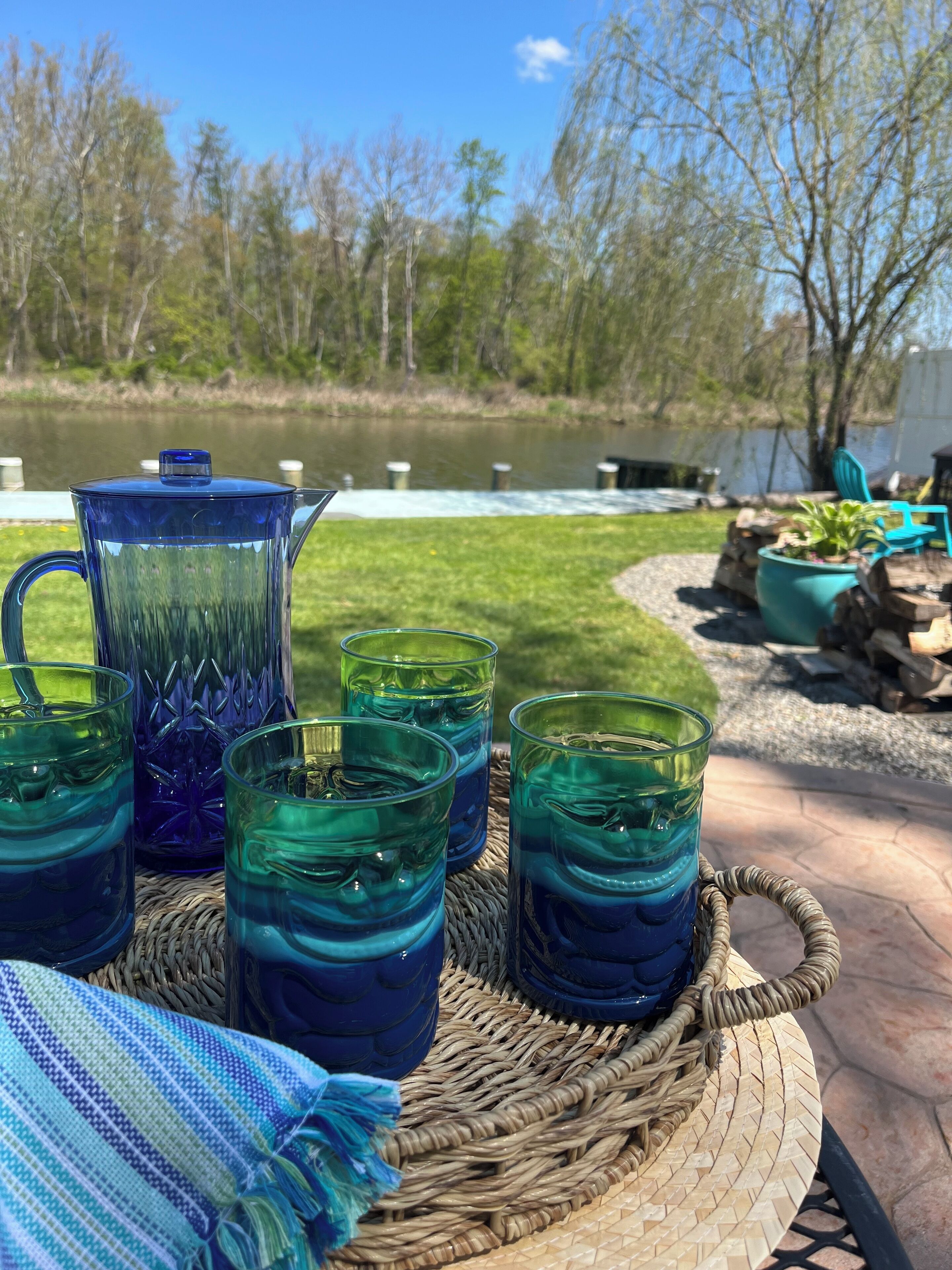 Ein Korbtisch mit einer klaren Glasgießkanne und vier Gläsern mit blauer Flüssigkeit, draußen mit Blick auf einen Fluss und Bäume.