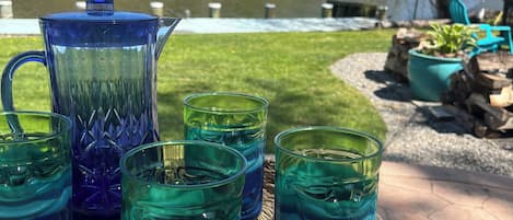 Ein Korbtisch mit einer klaren Glasgießkanne und vier Gläsern mit blauer Flüssigkeit, draußen mit Blick auf einen Fluss und Bäume.