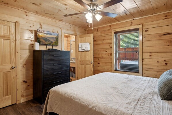 Un dormitorio de cabaña de madera con cama, cómoda, ventilador de techo y ventana con persianas.