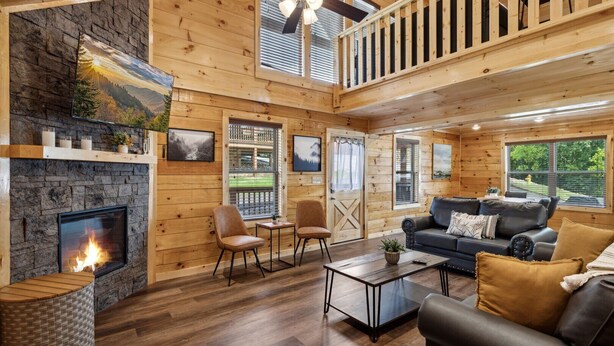 A cozy cabin interior with a stone fireplace, wooden walls, a balcony, and a comfortable seating area.