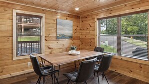 A wooden interior with a dining table, four chairs, a painting, and large windows.