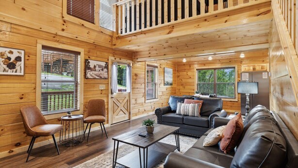 A cozy cabin interior with a wooden interior, a black leather sofa, a dining table, and a staircase leading to an upper level. - Amazing Grace - Newly Built - 3 BR - Less Than 2 Miles from The Island (Pigeon Forge)
