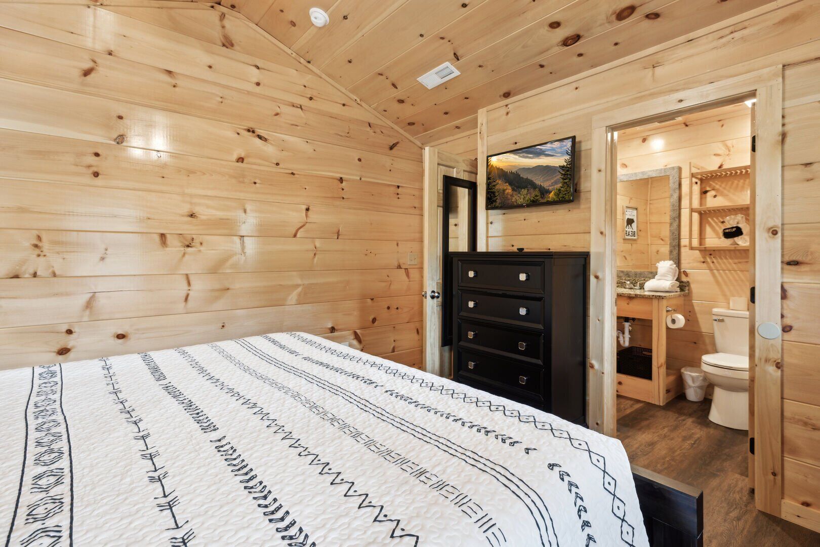 A wooden cabin room with a bed, dresser, and a bathroom visible through an open door.