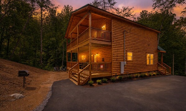 Une cabane en rondins à deux étages, dotée d’un balcon, d’un porche à l’avant et d’une allée menant à l’entrée.