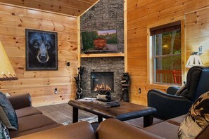 L’intérieur d’un chalet chaleureux, avec une cheminée en pierre, des murs en bois, un portrait de chien encadré et une fenêtre donnant sur la forêt.