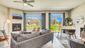 A modern living room with a fireplace, a sofa, a dining table, and large windows offering a view of the neighborhood. - Mid-Brambleberry (Colorado Springs)