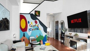A modern living room with a large colorful mural, a fireplace, and a Netflix TV. - Colorful Colorado house by DIA Airport Rockies & Downtown! (Commerce City)