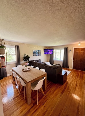 A spacious living area with a wooden dining table, white chairs, a dark sofa, and a TV mounted on the wall. - Casa Bemidji - 5 Beds, Prime Location, Spacious & Family Friendly (Bemidji)