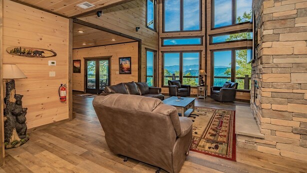 A spacious living room with a large window, wooden flooring, and a stone accent wall. - Mountain Dreams - Mountain views for miles! (Sevierville)