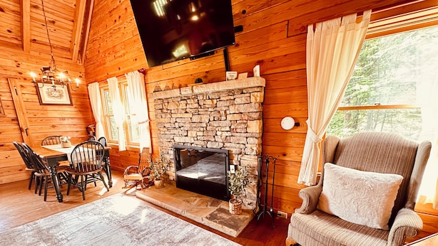 A cozy cabin interior with a stone fireplace, wooden walls, a dining area with chairs, and a television mounted on the wall.