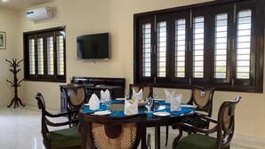 A dining area with a round table set for six, surrounded by wooden chairs with green cushions. There is a flat-screen TV mounted on the wall above the table. - The Udaisagar Retreat - Udaipur (Udaipur)